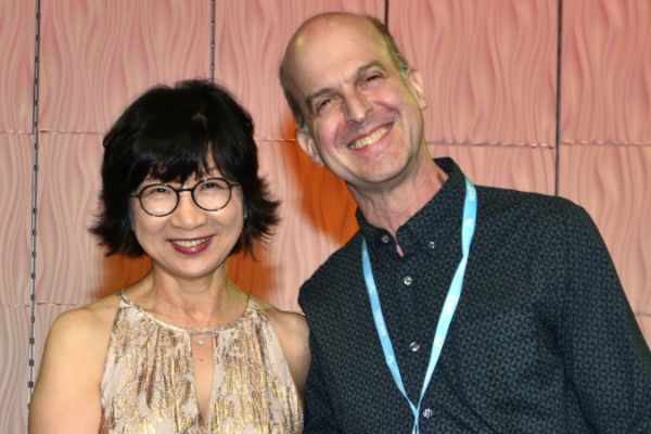 An Asian woman with short dark hair and glasses stands with a white man in a dark patterned shirt with short hair.