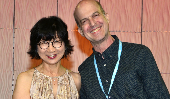 An Asian woman with short dark hair and glasses stands with a white man in a dark patterned shirt with short hair.
