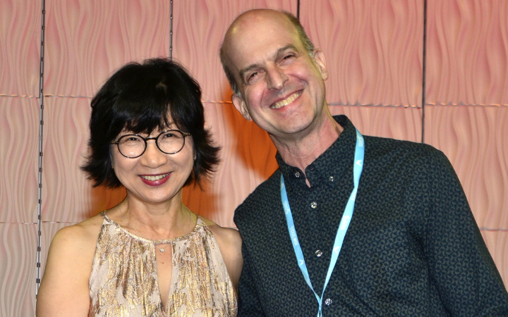 An Asian woman with short dark hair and glasses stands with a white man in a dark patterned shirt with short hair.