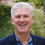 Rob Gould smiles at the camera. He is wearing a blue shirt and jacket and has short white hair.