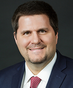 A white man in a blue suit jacket, white shirt, and red tie with short dark hair and a goatee.