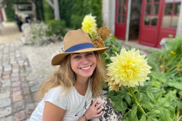 Long hair, big smile, wearing a hat and sitting next to flowers