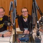 John (short hair, big smile) Rosemary, (hair pinned back, slight smile) and Richard Cambell (mustach and beard, big smile) sit around a desk with microphones in front of them