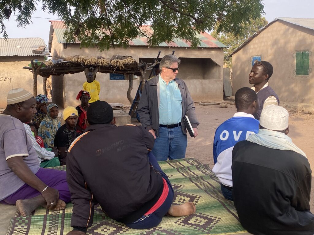 A white man and African man talk in front of a group of other Africans near a well.