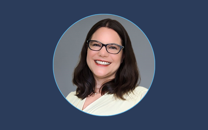 A woman with long dark hair smiles and is wearing glasses with dark rims.