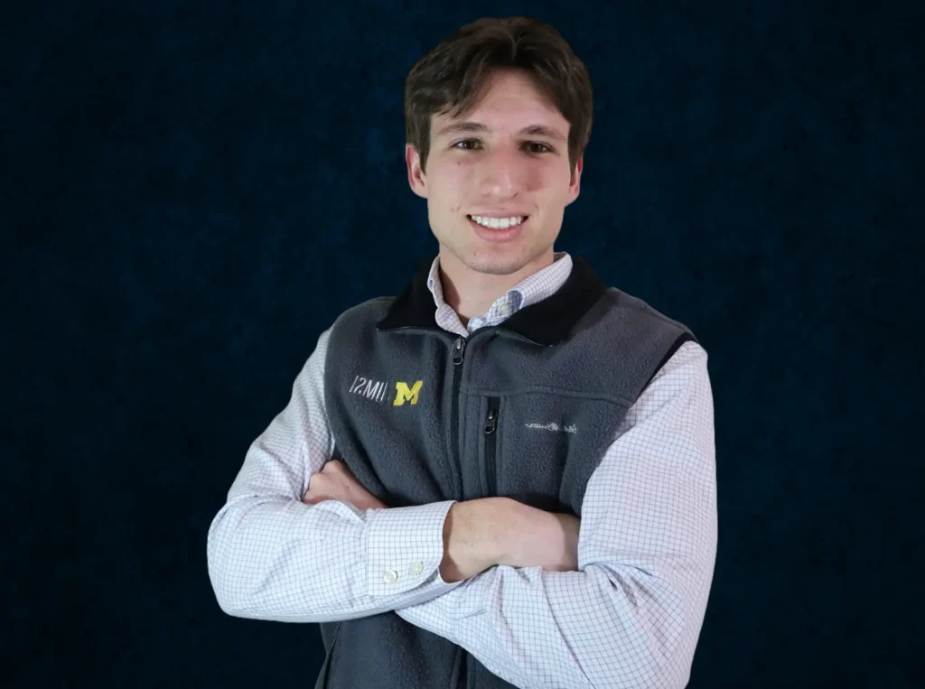 A photo of Avery, who has brown hair and is wearing a University of Michigan fleece vest and a light blue checkered shirt.