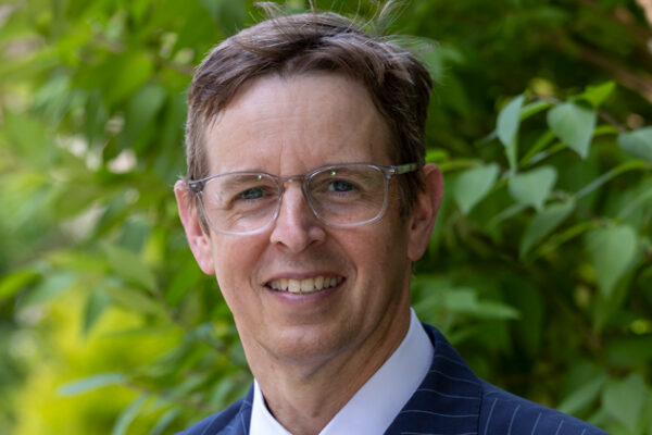 Photo of Greg, light brown hair, glasses, and a blue striped suit.