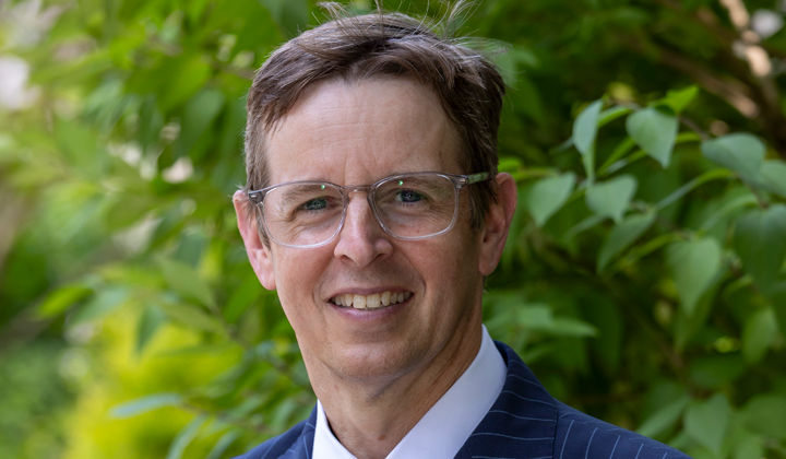 Photo of Greg, light brown hair, glasses, and a blue striped suit.