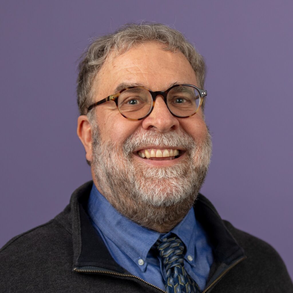 Nicholas Horton has short gray-brown hair, round glasses, and a beard, and is wearing a blue dress shirt with a black zip-up jacket.
