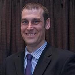 Justin Jacobs has short brown hair and is wearing a dark suit with a blue striped shirt.