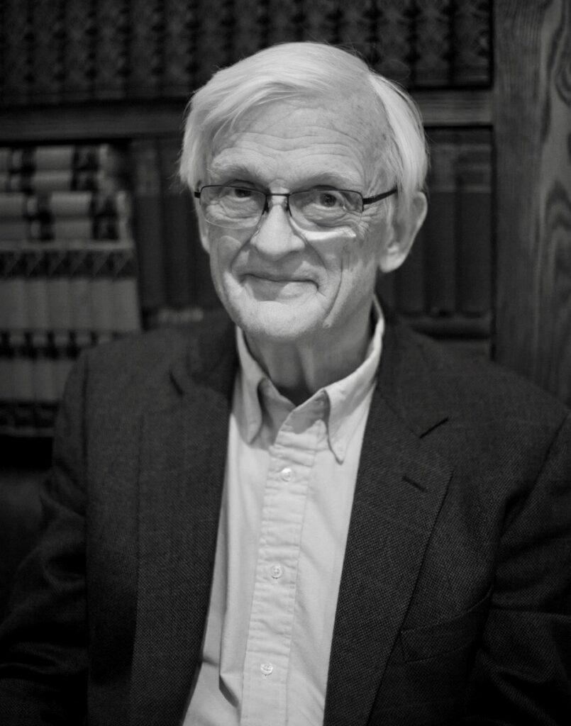 A black and white photo of Gudmund Ragnvaldsson Iversen with white hair and glasses, smiling at the camera in a light-colored dress shirt and dark suit jacket.