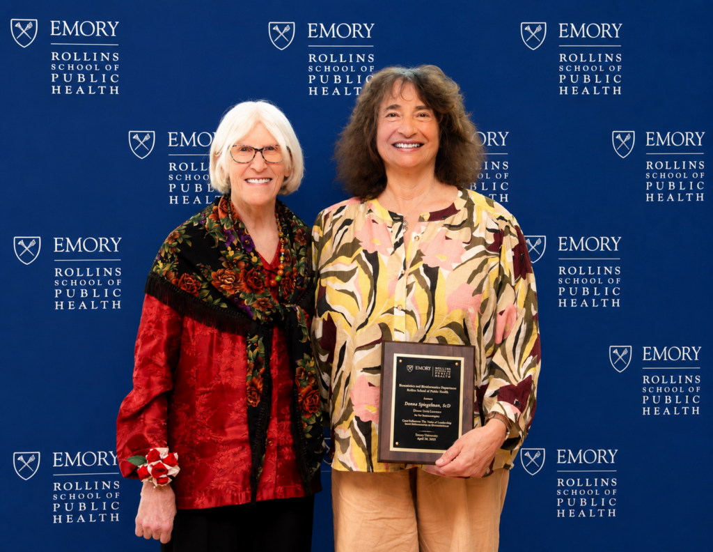 Donna Brogan (white hair and glasses in a red shirt) presents a plaque to Donna Spiegelman (curly brown hair, yellow patterned shirt) with a plaque in front of an Emory University Rollins School of Public Health background.