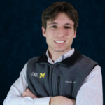 A photo of Avery, who has brown hair and is wearing a University of Michigan fleece vest and a light blue checkered shirt.