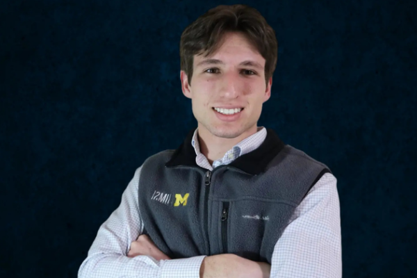 A photo of Avery, who has brown hair and is wearing a University of Michigan fleece vest and a light blue checkered shirt.