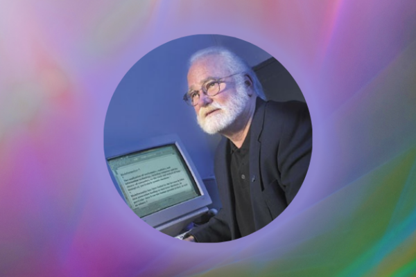 Karl Peace sits in front of a now-vintage computer, wearing a dark suit jacket and a white beard.