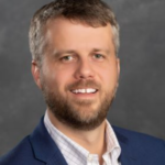 A white man with a beard wearing a white collared shirt and a navy jacket