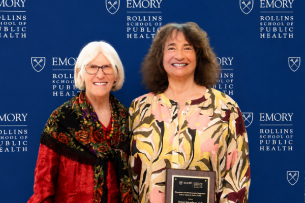 Donna Brogan (white hair and glasses in a red shirt) presents a plaque to Donna Spiegelman (curly brown hair, yellow patterned shirt) with a plaque in front of an Emory University Rollins School of Public Health background.
