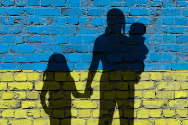 Silhouettes of a woman and two children against a brick wall painted blue and yellow, like the Ukrainian flag.