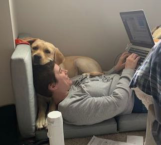 Erin Chapman works on a laptop while reclined. She has short brown hair and is wearing a gray sweatshirt. Her service dog, a yellow lab named Valor, sits next to her with his head resting on top of her head.