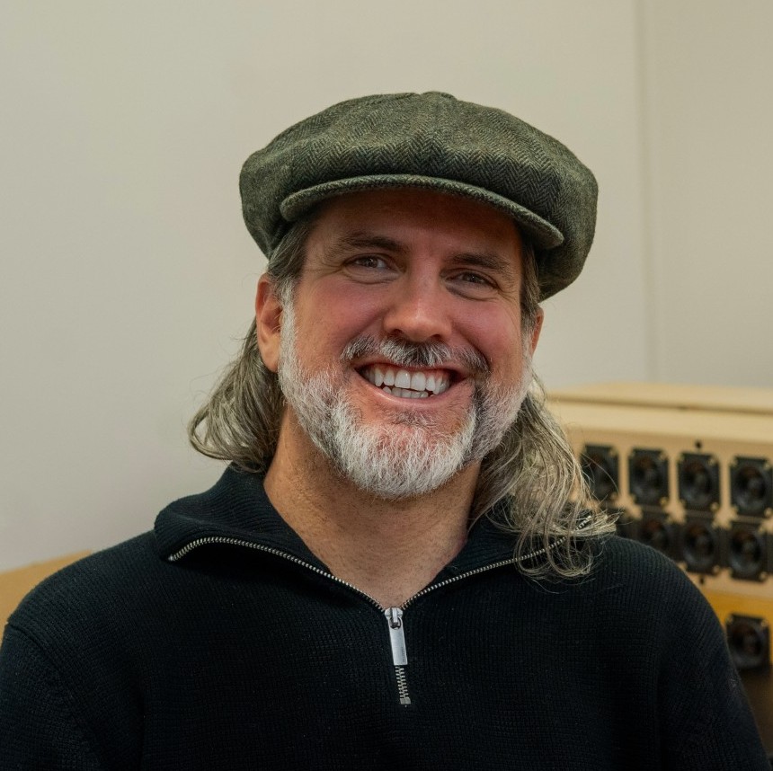 Kyle Keane smiles at the camera. He is wearing a black zip-up sweater and a dark olive green newsboy cap. He has shoulder-length gray hair and a gray beard.