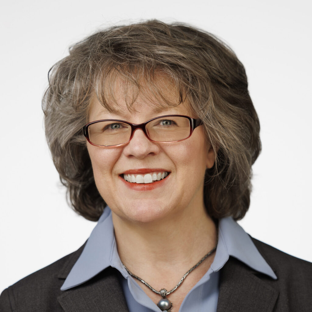 Rochelle Martinez wears a dark jacket, a blue collared shirt, glasses, and shoulder-length grayish-brown hair.