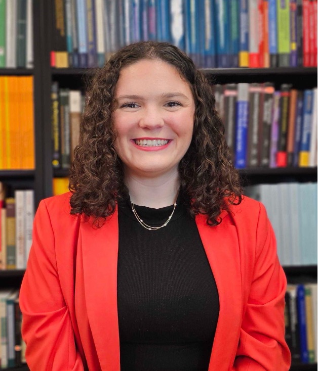 Aubree Krager has shoulder-length curly brown hair and is wearing a bright red jacket with a black shirt. She is smiling at the camera.