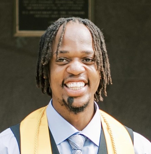Blessed Mbogo is wearing a light blue dress shirt and tie, and a yellow stole. He is smiling at the camera and has chin-length locs.