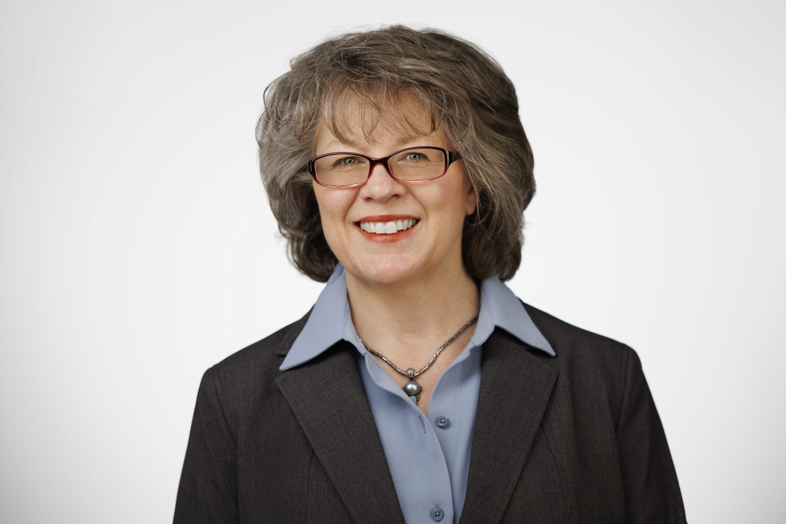 Rochelle Martinez wears a dark jacket, a blue collared shirt, glasses, and shoulder-length grayish-brown hair.