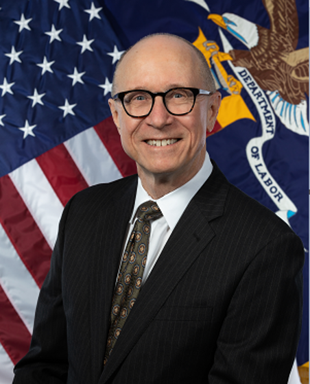 William Beach wears a dark suit and tie, glasses, and a bald haircut in front of an American Flag background.